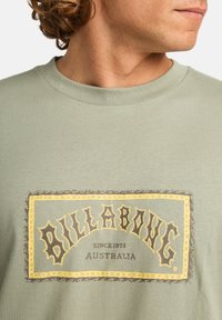 Camiseta de algodón verde oliva con un gran gráfico del logo amarillo y marrón con el texto "BILLABONG" y "DESDE 1973" en el centro.