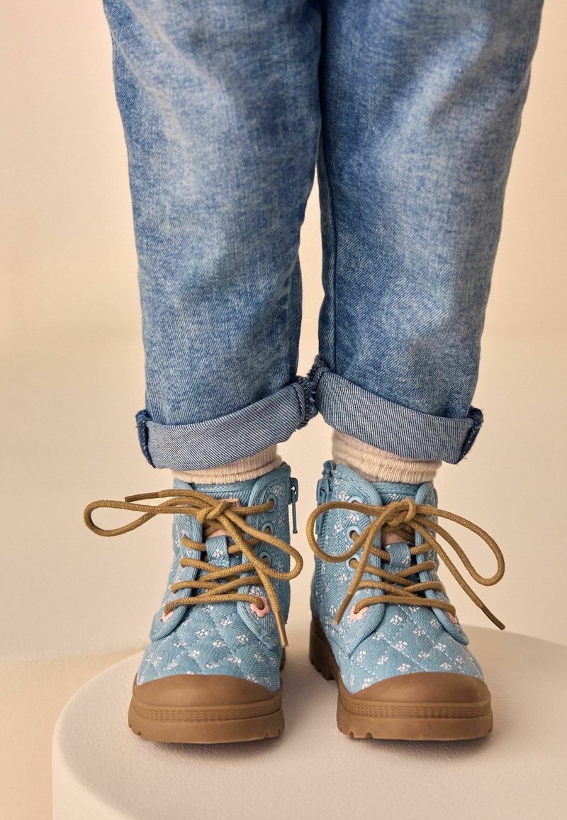 Enfant portant un jean bleu retroussé et des bottes bleu clair à motifs avec des semelles et lacets couleur beige, debout sur une plateforme blanche sur fond beige.
