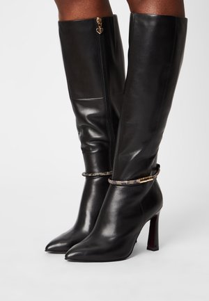 Bottes montantes en cuir noir avec bouts pointus, talons hauts, fermetures éclair latérales et brides de cheville décoratives ornées de strass portées aux jambes.