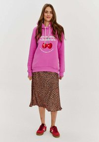 Ružová mikina s grafikou "Ochutnaj moje čerešne", spárovaná s midi sukňou s leopardím vzorom a červenými teniskami. Hladká látka a voľný strih.