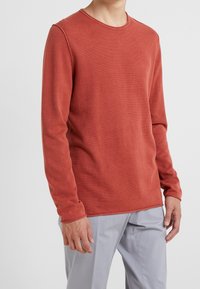 Långärmad, rostfärgad skjorta i texturerad tyg med rund hals och synliga sömdetaljer, kombinerad med ljusgrå byxor.