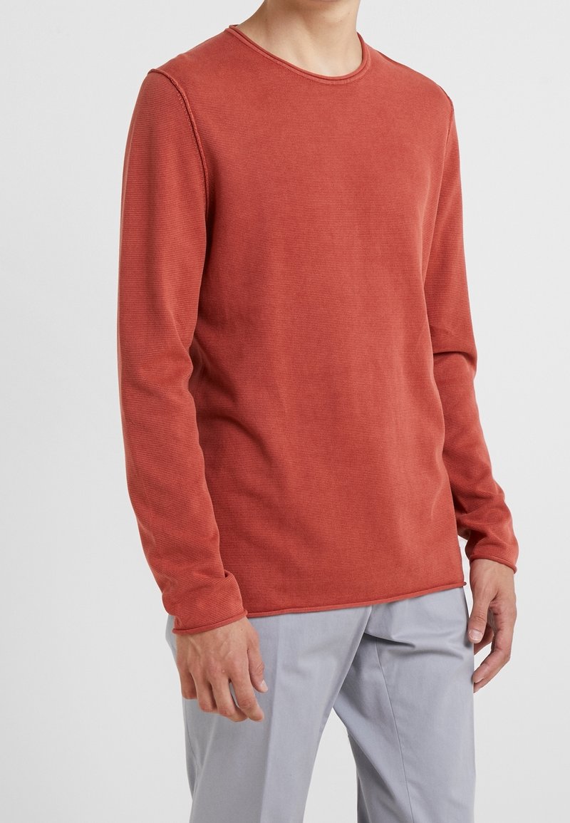 Långärmad, rostfärgad skjorta i texturerad tyg med rund hals och synliga sömdetaljer, kombinerad med ljusgrå byxor.