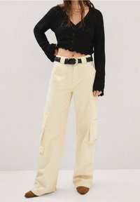 Zwarte gebreide cropped sweater met een V-hals, gecombineerd met crèmekleurige cargobroek, versierd met zijzakken en een gevlochten zwarte riem. Bruine puntige schoenen.