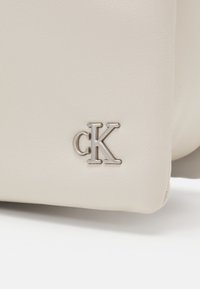 Borsa in ecopelle beige con texture liscia, caratterizzata da un logo "cK" argentato metallico sul davanti. Nessun ulteriore ornamento.