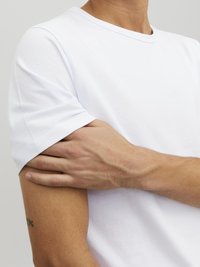 Bijela pamučna majica s okruglim izrezom i kratkim rukavima, prikazana izbliza s rukom koja počiva na bicepsu. Glatka tekstura.