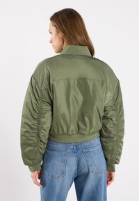 Maslinasto zelena bomber jakna s izrezanim dizajnom, nabrane bočne detalje i rebraste manžete, uparen s svijetloplavim trapericama.