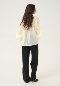 Blusa leggera gialla chiara, trasparente, con maniche a sbuffo e dettagli arricciati sulle spalle, abbinata a pantaloni neri a gamba larga e ballerine nere.