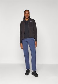 Giacca nera con zip, pantaloni blu e sneakers nere. La giacca ha una texture liscia con una tasca sul petto. I pantaloni sono tapered e realizzati in un tessuto leggero.