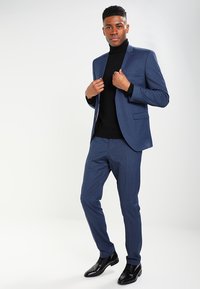 Marinblå kostym med slim-fit design, strukturerad tyg och matchande byxor. Bärs över en svart polotröja, ihop med glänsande svarta finskor.