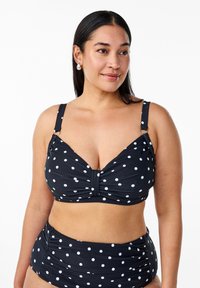 Juoda bikini komplektas su baltais taškeliais, sukurtas su vyniotu priekiu, reguliuojamomis petnešėlėmis ir aukštu liemeniu, kuriame yra šoninės raukšlės.