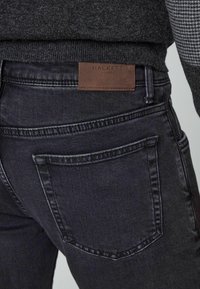 Vue arrière d'un jean en denim gris foncé avec une étiquette en cuir marron Hackett London sur la ceinture, porté avec un pull texturé gris.
