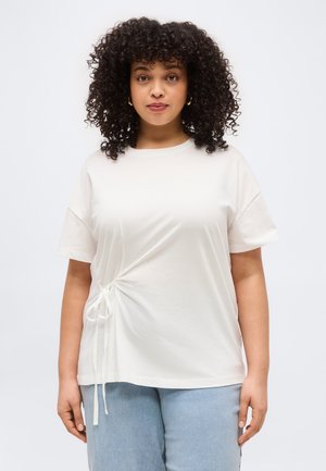 Nainen kiharatukkaisena, valkoinen lyhythihainen paita sivusolmulla ja vaaleansiniset farkut, seisomassa yksivärisen taustan edessä.