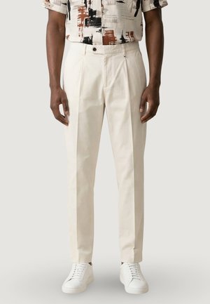 Mężczyzna w kremowych, dopasowanych spodniach, białych trampkach i koszuli z krótkim rękawem w abstrakcyjny wzór, stojący na tle jednolitego tła.