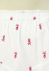 Tejido blanco con bordado floral rojo, textura ligera, cinturilla fruncida y delicados acentos de costura a lo largo del diseño.