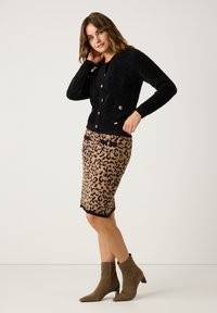 Frau in einem schwarzen, geknöpften, flauschigen Cardigan, einem Bleistiftrock mit Leopardenmuster und braunen Wildleder-Stiefeletten, die vor einem schlichten Hintergrund posiert.
