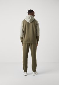 Completo sportivo verde oliva con giacca a cappuccio grigio chiaro. Presenta una vestibilità rilassata, polsini a coste e caviglie elasticizzate. Tessuto morbido.