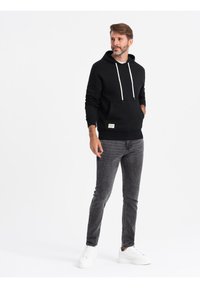 Sudadera negra con cordones blancos, bolsillo tipo canguro y detalle de etiqueta, combinada con jeans slim-fit gris oscuro y zapatillas blancas.