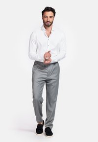 Witte blouse met lange mouwen en knopen, grijze op maat gemaakte broek en zwarte instapschoenen. De stof heeft een gladde afwerking; de algehele uitstraling is netjes.