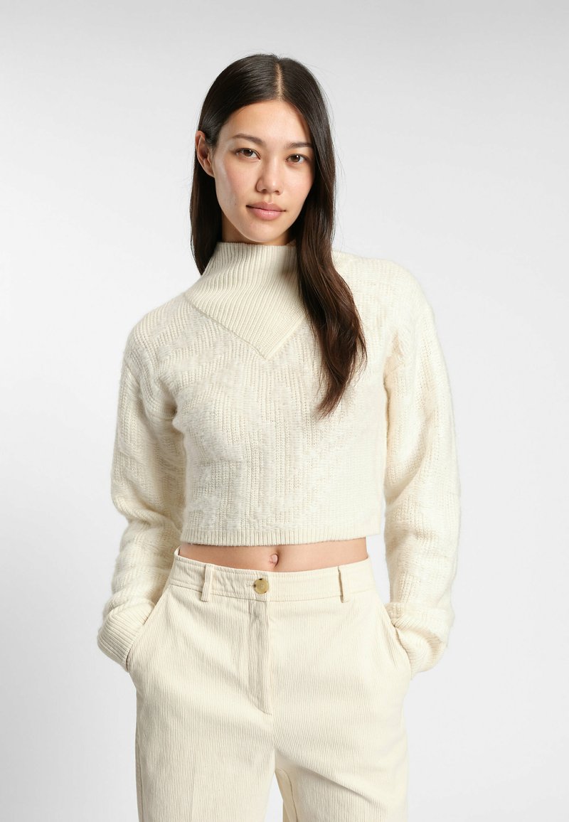 Cream-colored cropped sweater with a high collar and textured pattern. Paired with light beige corduroy pants that have a front button.