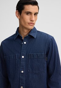 Donkerblauwe denim shirt met een knoopsluiting aan de voorkant, twee borstzakken en zilveren knopen. De stof heeft een gestructureerde, geweven uitstraling.