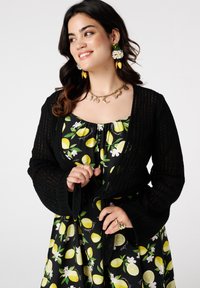 Een lachende vrouw draagt een zwarte cardigan over een bloemenjurk met citroenen. Ze draagt opvallende oorbellen en een ketting.