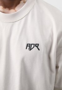Hellgraues T-Shirt mit kurzen Ärmeln, das auf der linken Seite ein schwarzes "ADR"-Logo trägt. Der Stoff wirkt weich mit einer glatten Textur.
