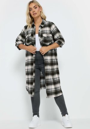 Giacca a camicia a quadri in bianco e nero con una vestibilità lunga e ampia, design con bottoni e maniche arrotolate; abbinata a pantaloni grigi e sneakers bianche.