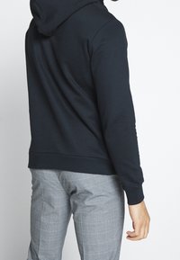 Felpa blu scuro con tasca a marsupio, polsini e orlo a costine. Indossata con pantaloni grigi a quadri, caratterizzati da una texture liscia.