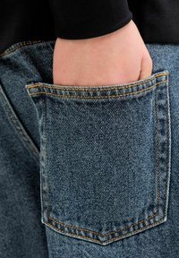 Kéz benyúlva a kék farmernadrág hátul zsebébe, látható sárga varrással, fekete felső ellen.