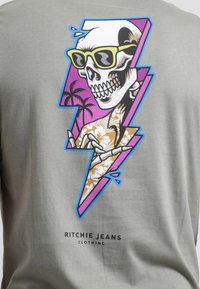 T-shirt gris clair avec un graphique coloré d'un crâne portant des lunettes de soleil, orné de motifs floraux et d'accents en éclairs. Logo de la marque en bas.