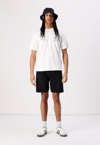 Tricou alb din bumbac cu imprimeu logo, pantaloni scurți din denim negri și pălărie tip bucket neagră. Pantofi sport gri și albi cu dungi și șosete tubulare.