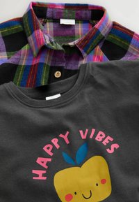 Chemise à carreaux en violet, vert et noir avec un col à boutons sur un t-shirt gris présentant une pomme souriante jaune et le texte "HAPPY VIBES" en rose.