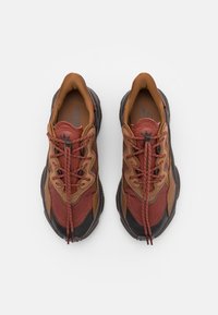 Zapatillas deportivas en tonos marrones y óxido, con parte superior de cuero texturizado y malla, que presentan suelas negras gruesas y cordones en contraste.