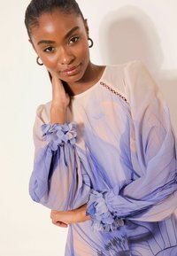 Gennemsigtig bluse med lange ærmer i bløde pastelfarver med blå og lilla abstrakte mønstre. Har teksturerede blomsterdetaljer på manchetterne.