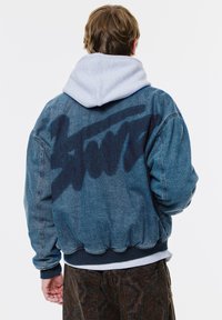 Jachetă din denim cu o glugă gri dedesubt. Logo întunecat albastru imprimat pe spate. Prezintă manșete și tiv cu zoom, croială lejeră.