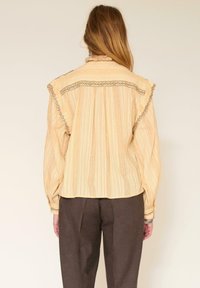 Blouse beige à rayures, ample, avec un col volanté et des coutures décoratives le long des épaules, associée à un pantalon marron foncé.