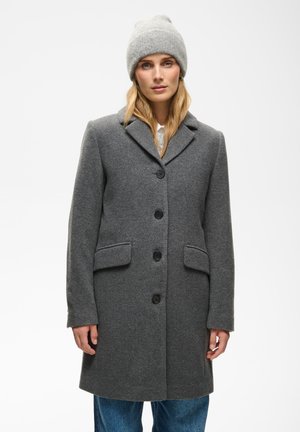 Femme portant un manteau en laine gris boutonné, un bonnet tricoté gris et un jean bleu, debout devant un fond clair uni.