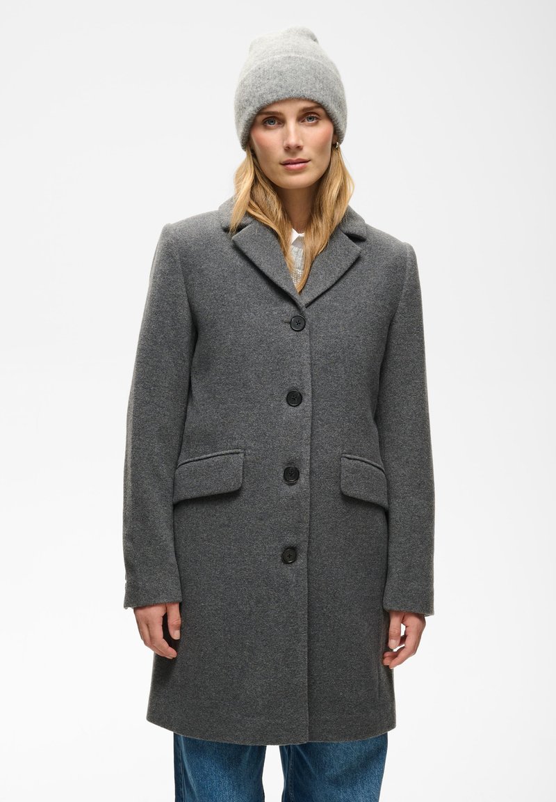Femme portant un manteau en laine gris boutonné, un bonnet tricoté gris et un jean bleu, debout devant un fond clair uni.