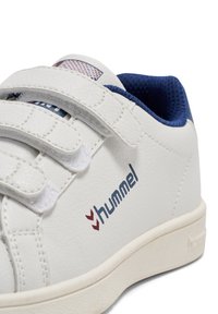 Biały syntetyczny sneaker z paskami na rzepy, granatową wyściółką, tłoczonymi detalami oraz logo Hummel w kolorze czerwonym. Zaokrąglony nosek i płaska podeszwa.
