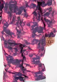 Barn iført en snedragt med pink og lilla galakse mønster, der har elastisk liv og manchetter, står med højre hånd synlig.