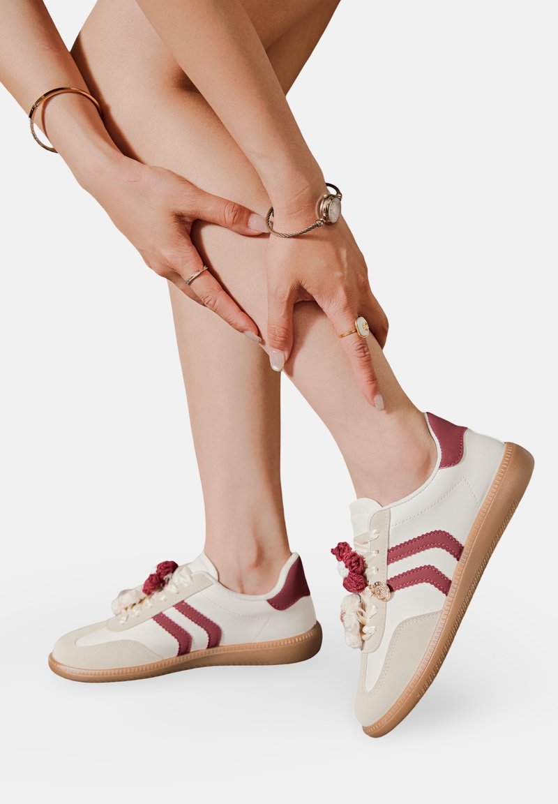Femme touchant sa jambe inférieure, portant des baskets blanches avec des rayures bordeaux et des lacets décoratifs en forme de rose sur un fond blanc.