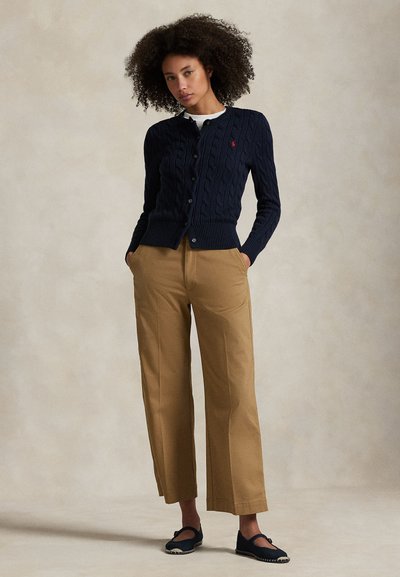 Göndör hajú nő, aki tengerészkék fonott mintás kardigánt, bézs bőszárú nadrágot és fekete Mary Jane cipőt visel, kezét zsebre dugva áll.