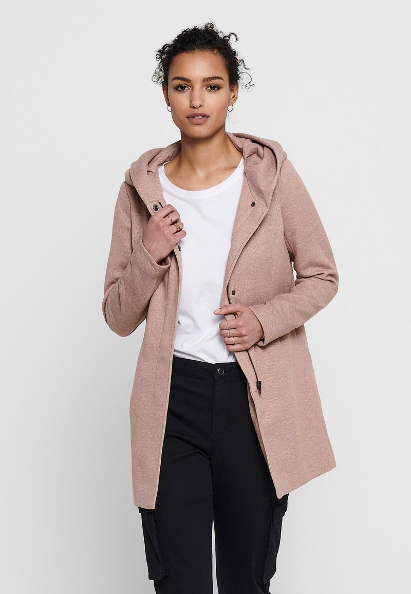 Cappotto cappuccio rosa chiaro con zip frontale, tessuto testurizzato e tasche laterali, indossato sopra una maglietta bianca e pantaloni neri.