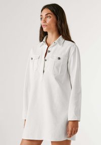 Abito bianco a maniche lunghe in cotone con collo a camicia, patta con bottoni e due tasche sul petto. Presenta una vestibilità comoda e una texture morbida.