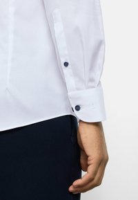 Weißes strukturiertes Hemd mit spitzem Kragen, Knopfmanschetten mit marineblauen Knöpfen und tailliertem Schnitt. Kombiniert mit dunkler Hose.