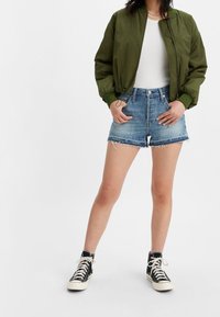 Olive green bomber jacket, white tank top, blue denim shorts with frayed edges, and black high-top sneakers with white soles.