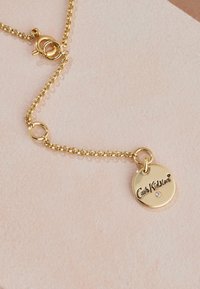 Zelta ķēdes kaklarota ar apaļu piekariņu, uz kura iegravēts "Cath Kidston" un neliels dārgakmens akcenti, izvietots uz teksturēta fona.