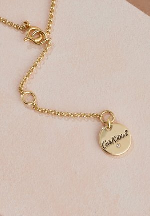 Guldkedja med runt hänge som har "Cath Kidston" ingraverat och en liten ädelstensaccent mot en strukturerad bakgrund.