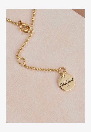 Kuldne kett-kaelakee, millel on ümmargune ripats "Cath Kidston" graveeringuga ja väike vääriskivi aksent, tekstuurse tausta peal.