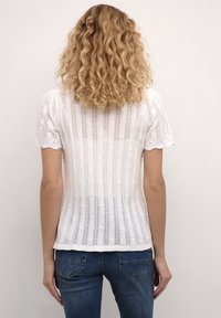 Vit stickad topp med vertikala ränder och en strukturerad mönstring, med korta puffärmar och scallopade kanter, som bärs med blå denimjeans.